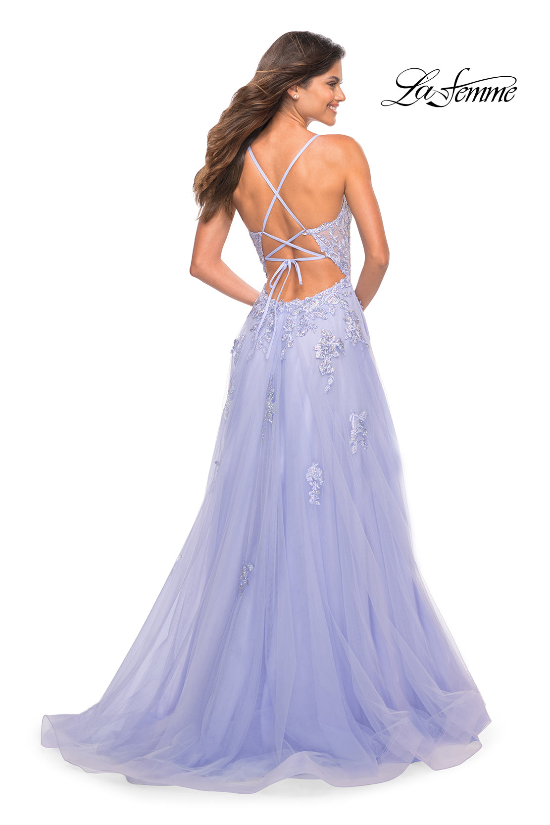 A woman stands facing away from the camera in a La Femme Open-Back Long Ball Gown with lace details and a strappy back, showcasing its elegant lavender design.